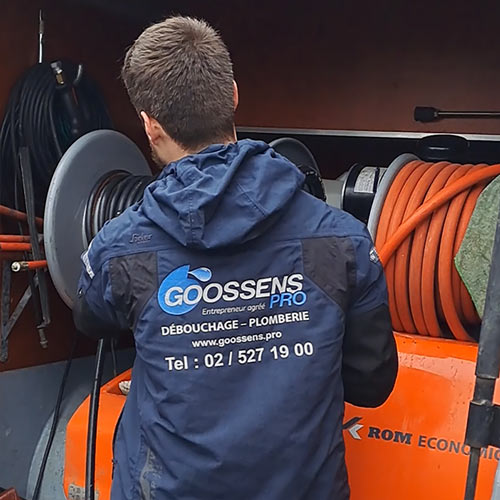 Technicien de GOOSSENS PRO effectuant un service de débouchage de canalisation avec un enrouleur de tuyau, vêtu d'une veste marquée 'GOOSSENS PRO Débouchage - Plomberie' à l'arrière.